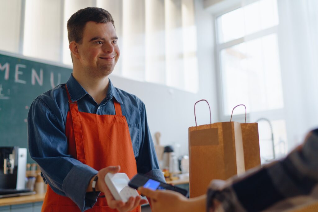 Cheerful young Down Syndrome waiter taking contactless smartphone payment from costumer in take away