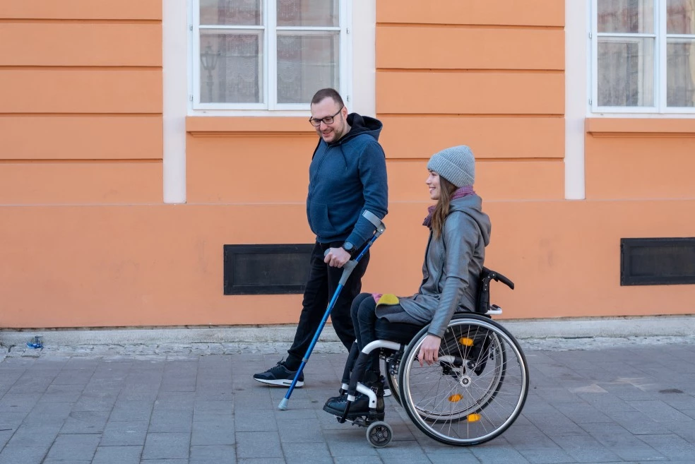 Two disabled friends, one in a wheelchair, walking down a street together and smiling